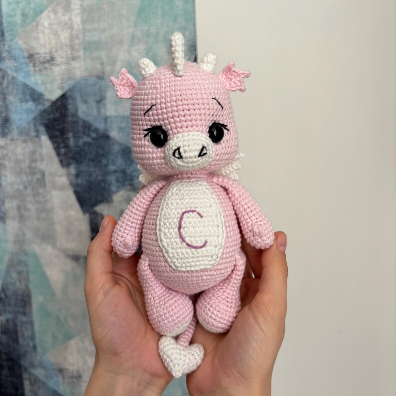 May include: A handmade pink crochet dragon doll with white accents, including wings, horns, and a heart-shaped tail. The dragon has a white oval on its chest with the letter "C". The doll is held in two hands.