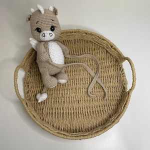 May include: A handmade crochet toy in the shape of a light brown and white mythical creature with horns, wings, and a heart-shaped tail. The toy is placed on a round, woven tray with handles. The background is white.