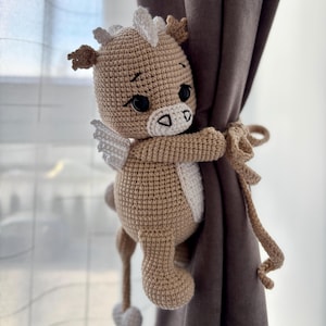 May include: A crocheted, light brown Pegasus-like creature with white wings and accents, holding onto a brown curtain. The handmade plushie has black eyes and a white belly. The curtain tieback is made of the same yarn.