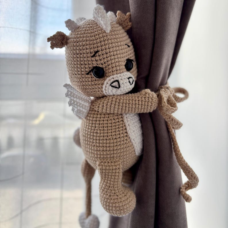 May include: A crocheted, light brown Pegasus-like creature with white wings and accents, holding onto a brown curtain. The handmade plushie has black eyes and a white belly. The curtain tieback is made of the same yarn.