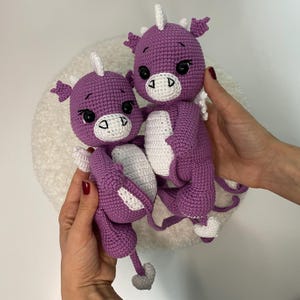 May include: Two handmade crochet dragon dolls. The dragons are purple with white accents on their bellies, horns, and feet. They have black eyes and are being held up by a person.
