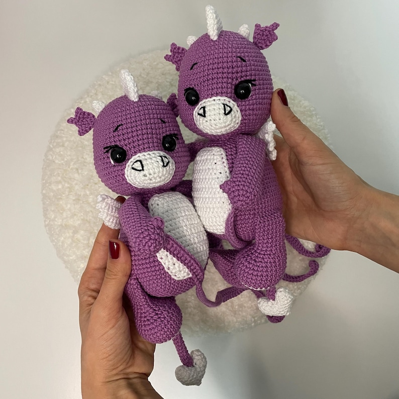 May include: Two handmade crochet dragon dolls. The dragons are purple with white accents on their bellies, horns, and feet. They have black eyes and are being held up by a person.