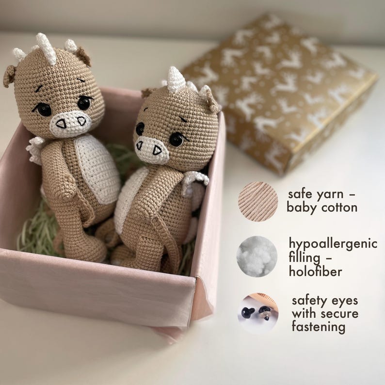 May include: Two handmade crochet dragon dolls in a pink box. The dragons are beige with white accents and black safety eyes. The image also shows text that says "safe yarn - baby cotton", "hypoallergenic filling - holofiber", and "safety eyes with secure fastening".