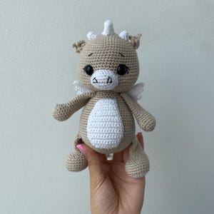 May include: A crocheted dragon plushie in tan and white. The handmade toy has large black eyes, small white wings, and a white belly. The dragon has a small horn and ear details. The plushie is held in a hand.