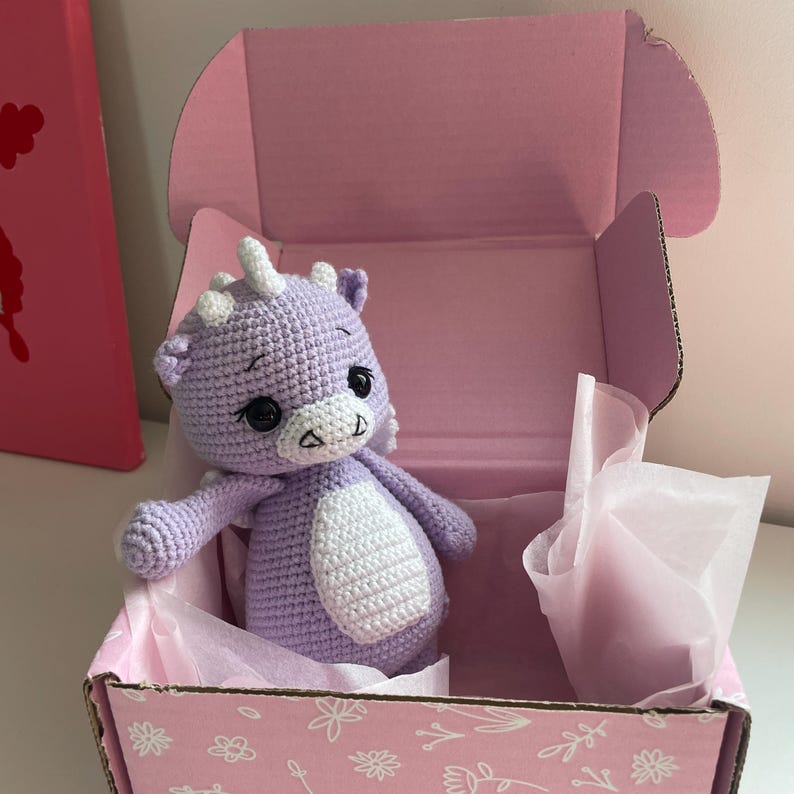 May include: A handmade, lavender crochet dragon toy with white accents, including a belly, horns, and facial details. The dragon is presented inside a pink box with floral designs, nestled on pink tissue paper.