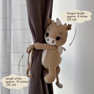 May include: A crocheted dragon curtain tieback in tan and white, approximately 8 inches (20 cm) tall. The ties are about 10 inches (26 cm) long. The dragon has black eyes and small white wings. The text on the image says "Dragon height - approx. 8 inches (20 cm)" and "Length of ties - approx. 10 inches (26 cm)".