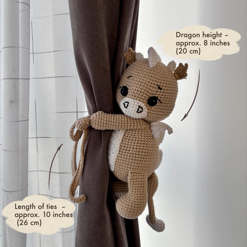 May include: A crocheted dragon curtain tieback in tan and white, approximately 8 inches (20 cm) tall. The ties are about 10 inches (26 cm) long. The dragon has black eyes and small white wings. The text on the image says "Dragon height - approx. 8 inches (20 cm)" and "Length of ties - approx. 10 inches (26 cm)".