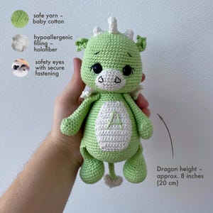 May include: A handmade green and white crocheted dragon toy. The dragon has black safety eyes, a white belly with the letter "A", and is approximately 8 inches tall. The text in the image indicates the toy is made with baby cotton yarn and hypoallergenic filling.