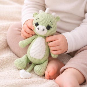 May include: A handmade green crochet dragon toy with white accents. The dragon has black eyes, a white belly, and small horns. The toy is held by a person, showcasing its soft texture and whimsical design. A charming, cuddly toy.