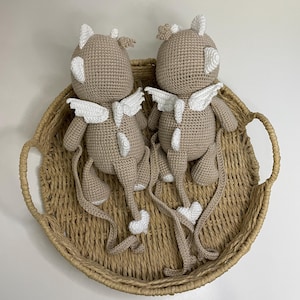 May include: Two crocheted plush toys in a light brown color, each with white wings, horns, and accents. The toys are placed in a woven basket with handles. The toys have long tails with heart-shaped details.