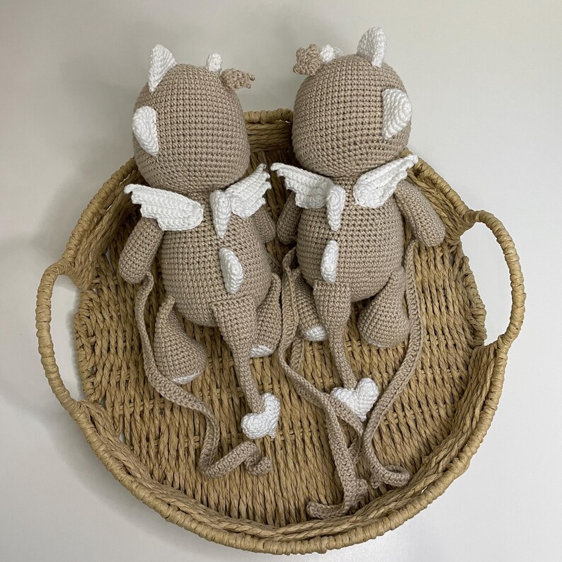 May include: Two crocheted plush toys in a light brown color, each with white wings, horns, and accents. The toys are placed in a woven basket with handles. The toys have long tails with heart-shaped details.