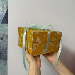 May include: A mustard-yellow gift box with a white leaf pattern and a light blue ribbon. The box is rectangular and held by hands. The ribbon is tied around the box and forms a bow.
