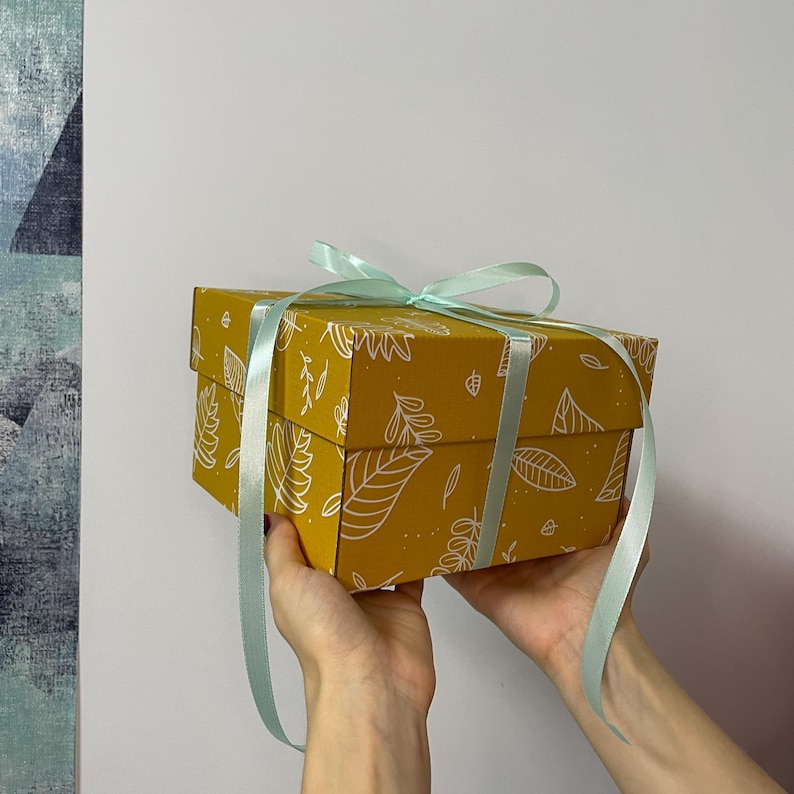 May include: A mustard-yellow gift box with a white leaf pattern and a light blue ribbon. The box is rectangular and held by hands. The ribbon is tied around the box and forms a bow.
