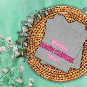 May include: A gray t-shirt with the words "OFFICIAL BARN CHORES SHIRT" in pink, folded on a woven wicker mat. The mat is on a light green surface, with sprigs of greenery and flowers around the edges.