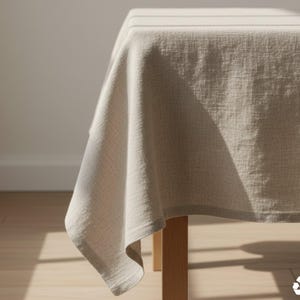 May include: A beige linen tablecloth draped over a wooden table. The fabric has a subtle woven texture and hangs in soft folds. The table is in a well-lit room, with sunlight casting shadows on the floor and wall. The tablecloth is made of 100% cotton.