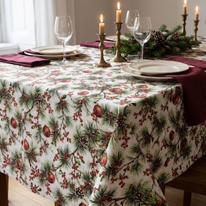 May include: A white tablecloth featuring a repeating pattern of green pine branches, brown pinecones, and red berries. The table is set with white plates, clear wine glasses, dark red cloth napkins, and brass candlesticks with lit candles.