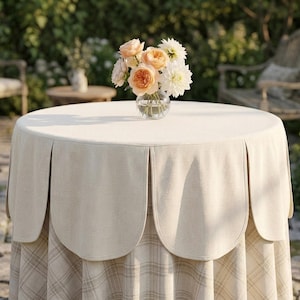 May include: A round table covered with a beige tablecloth featuring a scalloped edge overlay and a light tan plaid pattern underneath. A clear glass vase sits in the center, holding a bouquet of peach, pink, and white flowers.