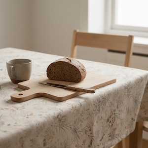 Puede incluir: Una escena de cocina r&uacute;stica con una tabla de cortar de madera, una hogaza de pan y un cuchillo. Una taza de cer&aacute;mica moteada est&aacute; cerca. La mesa est&aacute; cubierta con un mantel con estampado floral.