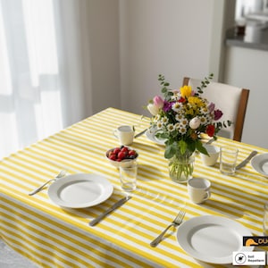 May include: A dining table set for a meal, featuring a yellow and white striped tablecloth. The table is set with white plates, cups, glasses, silverware, and a vase of colorful flowers. A bowl of berries is also present.