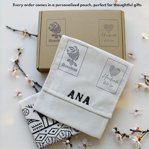 Puede incluir: Una caja beige y dos bolsas blanquecinas con texto y gr&aacute;ficos negros. La caja y las bolsas tienen un logotipo de rosa y coraz&oacute;n. Una bolsa tiene el nombre "ANA" impreso. El texto "Every order comes in a personalized pouch" es visible.