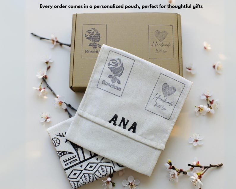 Puede incluir: Una caja beige y dos bolsas blanquecinas con texto y gr&aacute;ficos negros. La caja y las bolsas tienen un logotipo de rosa y coraz&oacute;n. Una bolsa tiene el nombre "ANA" impreso. El texto "Every order comes in a personalized pouch" es visible.