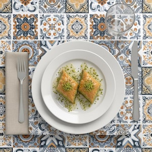 May include: Overhead shot of a table setting featuring two pieces of baklava on a white plate. The dessert is topped with chopped pistachios and sits on a patterned tablecloth with a fork, knife, and glass.