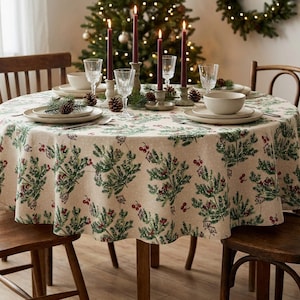 May include: A round beige tablecloth featuring a repeating pattern of green pine branches and red berries. The table is set with white ceramic plates, bowls, clear glass goblets, and four dark red taper candles in grey candle holders.