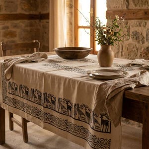 May include: A rustic dining table set with a beige tablecloth featuring a black animal and geometric pattern. The table is set with plates, napkins, a wooden bowl, and a vase with flowers. The room has a stone wall and a wooden chair.