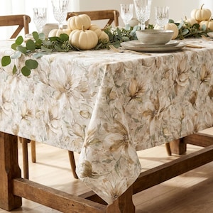 May include: A rectangular tablecloth featuring a watercolor-style floral pattern in shades of beige, brown, and muted green. The fabric drapes over a wooden table, which is set with white pumpkins, green eucalyptus sprigs, and ceramic dinnerware.