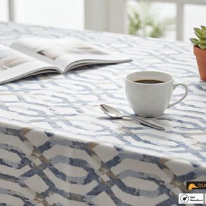 May include: A white tablecloth with a blue and gray geometric pattern. A white coffee cup with coffee, a spoon, an open book, and a potted succulent are on the table. The tablecloth is soil and water repellent.