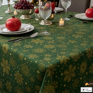 May include: A green tablecloth with a gold snowflake pattern, set for a festive occasion. The table is set with white plates, silverware, and crystal glassware. Red pomegranates and grapes are in silver bowls.