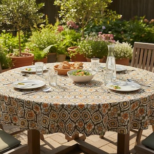 Può includere: Un tavolo rotondo apparecchiato per un pasto all'aperto. Il tavolo è coperto da una tovaglia fantasia nei toni del marrone, arancione e bianco. Il tavolo è apparecchiato con piatti, bicchieri, posate e una ciotola di insalata. Sono presenti anche un cestino di pane e una caraffa d'acqua.