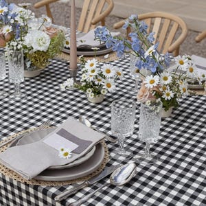 May include: A beautifully set table with a black and white checkered tablecloth. The table is adorned with floral arrangements of daisies and other flowers, crystal glasses, and place settings with linen napkins. Wooden chairs are in the background.