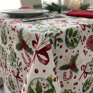 May include: A white tablecloth featuring a festive holiday pattern of red ribbons, green holly leaves, red berries, candy canes, and round ornaments. The fabric is draped over a table, with a red napkin and white plates visible on the surface.