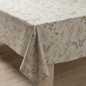Puede incluir: Un mantel rectangular con un estampado floral. La tela es de color beige claro con un dise&ntilde;o bot&aacute;nico en tonos grises, verdes y naranjas. El mantel est&aacute; drapeado sobre una mesa, mostrando el patr&oacute;n y la textura.