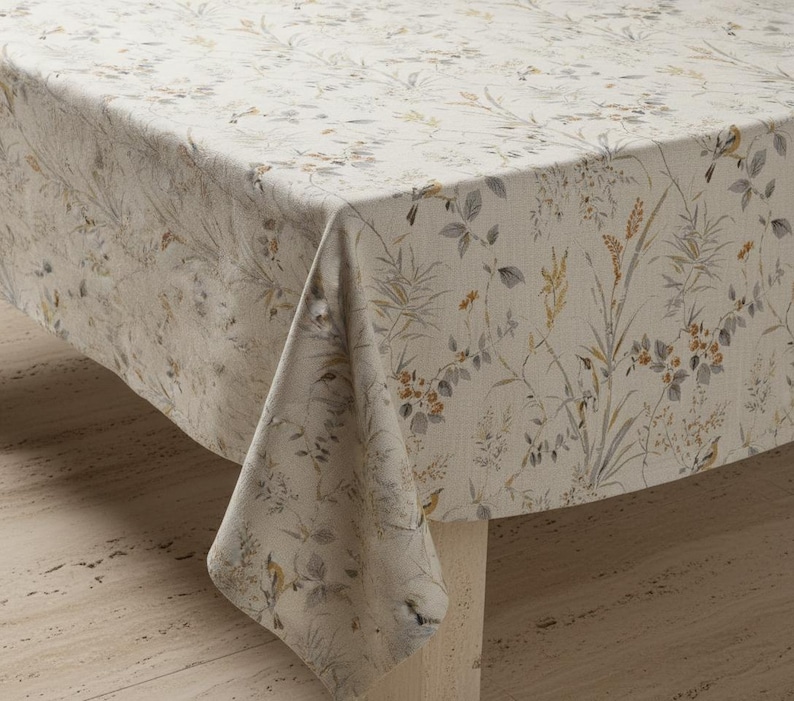 Puede incluir: Un mantel rectangular con un estampado floral. La tela es de color beige claro con un dise&ntilde;o bot&aacute;nico en tonos grises, verdes y naranjas. El mantel est&aacute; drapeado sobre una mesa, mostrando el patr&oacute;n y la textura.