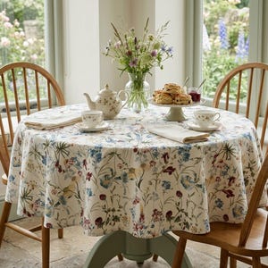 Peut inclure: Une table à manger ronde recouverte d'une nappe crème ornée d'un imprimé botanique coloré de fleurs sauvages. La table est dressée avec une théière à motifs floraux, deux tasses assorties, deux serviettes pliées, un vase et des scones.