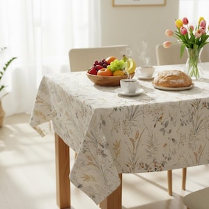 Puede incluir: Un mantel rectangular con un estampado floral cubre una mesa de madera preparada para una comida. Un cuenco de madera con fruta, tazas de caf&eacute;, pan y un jarr&oacute;n con tulipanes est&aacute;n sobre la mesa. El mantel tiene un fondo beige con dise&ntilde;os florales verdes y amarillos.