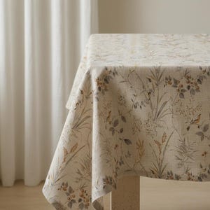 May include: A beige tablecloth with a botanical print featuring leaves, flowers, and birds in shades of brown, gray, and green. The tablecloth is draped over a table, showcasing the detailed pattern and texture of the fabric.