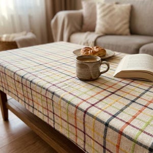 Peut inclure: Une nappe à carreaux beiges recouvre une table basse en bois. Une tasse en céramique avec du café, une assiette avec une pâtisserie et un livre ouvert reposent sur la table. Le motif à carreaux comprend des lignes rouges, jaunes, vertes et bleues.
