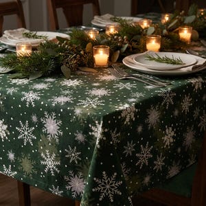 May include: A dark green tablecloth featuring a repeating pattern of white and light grey snowflakes of various sizes. The fabric is draped over a dining table set with white plates, silver forks, and small glass candle holders with lit candles.