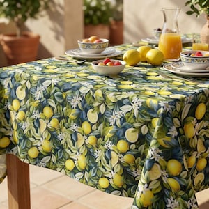 Pode incluir: Uma toalha de mesa retangular com um padrão vibrante de limões e folhas. O design apresenta limões amarelos, flores brancas e folhas verdes sobre um fundo azul escuro. A mesa está posta com taças de fruta, pratos e uma jarra de sumo.