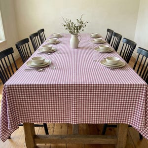 Puede incluir: Una mesa de comedor rectangular larga cubierta con un mantel de cuadros vichy rosas y blancos. La mesa está puesta con diez cuencos de cerámica blanca sobre platos a juego, dispuestos en parejas. Una jarra blanca con vegetación está en el centro.