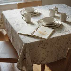 Puede incluir: Una mesa de comedor con un mantel floral, cuencos, platos, tazas y un cuaderno de cer&aacute;mica blanca. La mesa est&aacute; puesta con un mantel crema con un dise&ntilde;o bot&aacute;nico y sillas de madera.