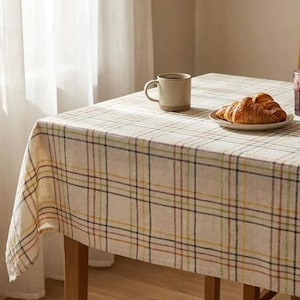 May include: A rectangular tablecloth featuring a cream-colored base with a multi-colored plaid pattern in thin lines of yellow, blue, and maroon. The fabric drapes over the edges of a wooden table, which holds a ceramic mug and a small plate of croissants.