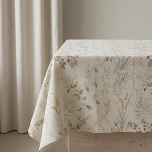 Puede incluir: Un mantel color crema con un estampado bot&aacute;nico que presenta p&aacute;jaros, hojas y hierbas en tonos grises, amarillos y verdes. La tela est&aacute; drapeada sobre una mesa, mostrando el dise&ntilde;o floral detallado. El mantel a&ntilde;ade un toque de elegancia inspirada en la naturaleza.