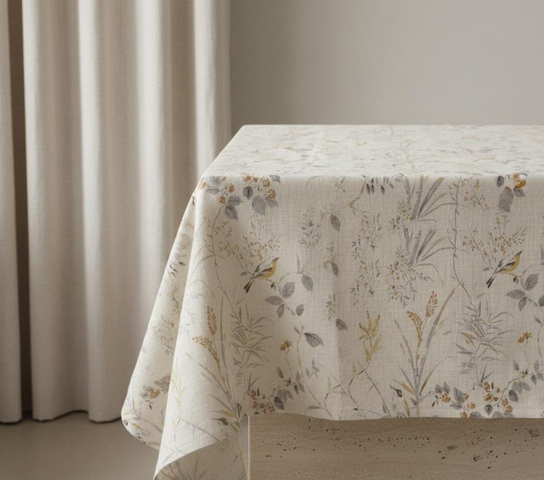 Puede incluir: Un mantel color crema con un estampado bot&aacute;nico que presenta p&aacute;jaros, hojas y hierbas en tonos grises, amarillos y verdes. La tela est&aacute; drapeada sobre una mesa, mostrando el dise&ntilde;o floral detallado. El mantel a&ntilde;ade un toque de elegancia inspirada en la naturaleza.