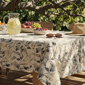 May include: An outdoor table set with a floral tablecloth, a jug of lemonade, bowls of fruit, sunglasses, and a straw hat. The tablecloth has a beige background with grey and brown leaf patterns.