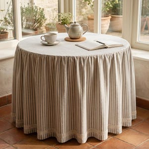 May include: A round table covered with a floor-length, beige and white striped tablecloth featuring a gathered skirt and a decorative lace trim along the bottom edge. On the table sits a ceramic teapot, a matching teacup, and an open book.