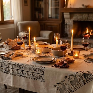 May include: A dining table set for a meal, featuring a white tablecloth with a black pattern. The table is set with plates, glasses of red wine, napkins, and lit candles. A fireplace and chairs are in the background.